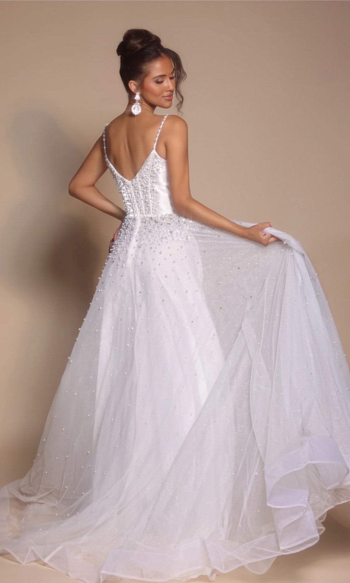 A woman in the Corseted Ball Gown: Portia and Scarlett PS26208, a white beaded sleeveless dress with a tulle skirt and long train, stands facing away and looks over her shoulder against a beige background.